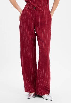 Femme portant un pantalon bordeaux taille haute à jambes larges avec des fines rayures blanches et des chaussures pointues argentées sur fond blanc.