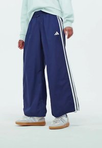 Marineblaue weit geschnittene Hose aus leichtem Material, mit weißen seitlichen Streifen und gesticktem Logo. Getragen mit weißen Schuhen mit Gummisohle.
