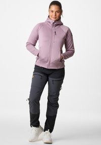 Lichtpaarse ritsjack met capuchon, voorzien van een zak op de borst, gecombineerd met zwarte wandelschoenen en witte sportieve schoenen.