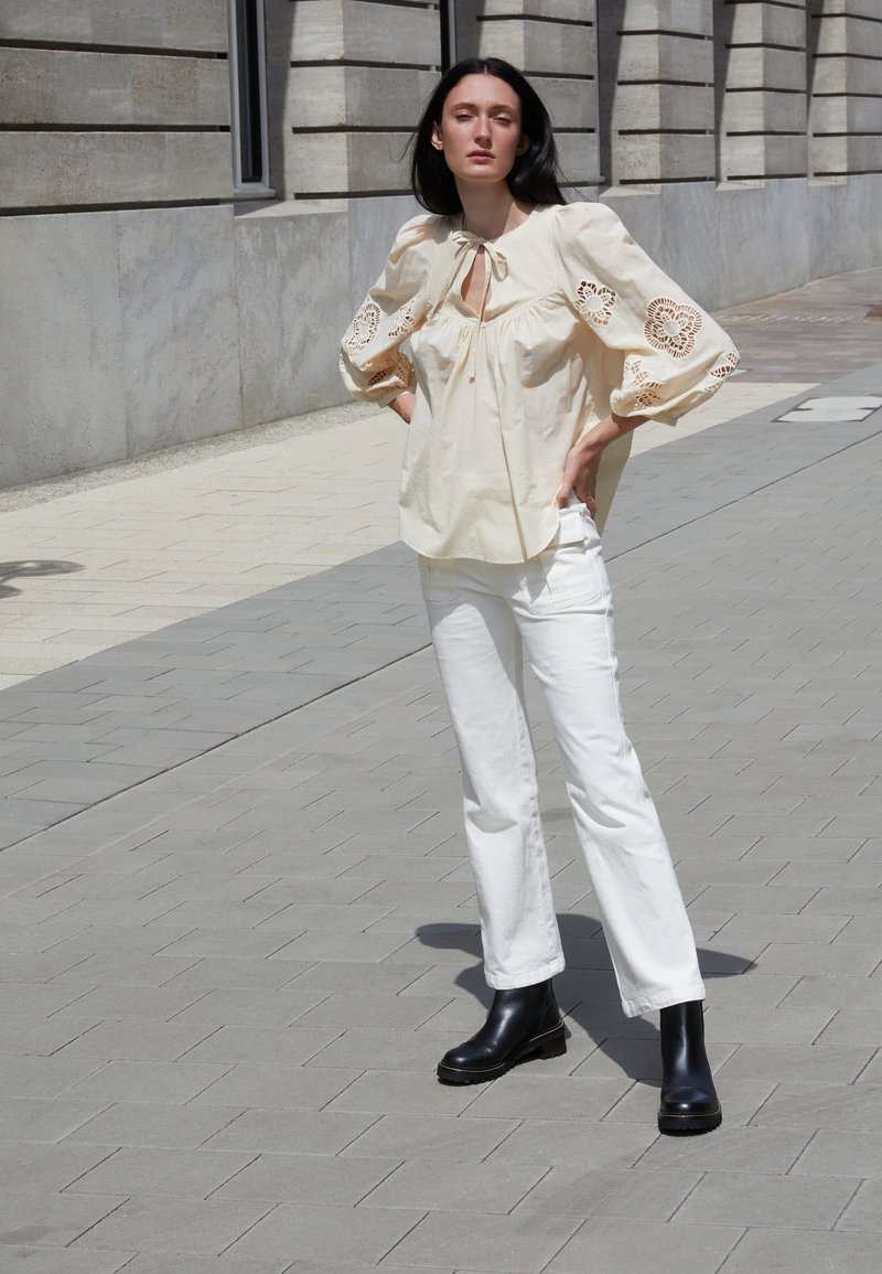 Blouse crème avec des manches bouffantes, agrémentée d'empiècements en dentelle. Associée à un pantalon droit blanc et des bottines noires.