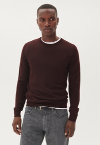 Burgundy stickad tröja med rund halsringning och ribbad mudd, parat med grå jeans. Slät textur, minimal detaljering.