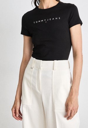 Woman wearing a black fitted T-shirt with "TOMMY JEANS" in white letters and high-waisted white pants with large front pockets.