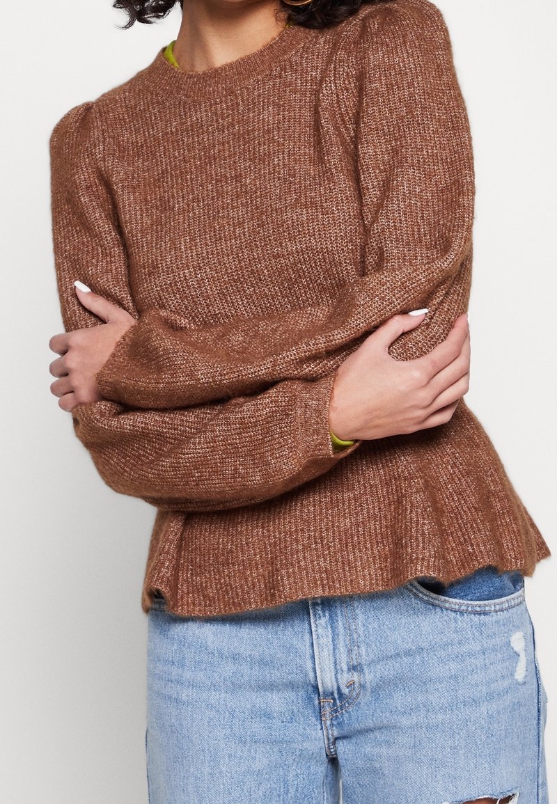 Personne portant un pull en tricot marron à manches bouffantes, les bras croisés, associé à un jean en denim bleu clair déchiré.