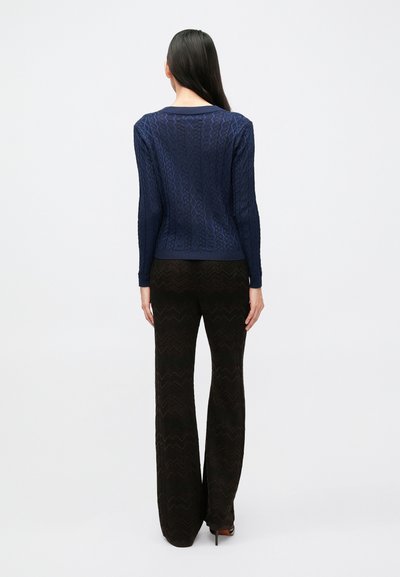 Tamsiai mėlynas megztinis su pynimo raštu ir ilgomis rankovėmis, derintas su juodomis platėjomis kelnėmis su zigzago raštu. Megztinis turi briaunotą apykaklę ir rankogalius.