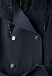 Gros plan d'un manteau foncé avec des boutons en métal étiquetés "CECIL" et des cordons noirs embossés avec "EVERYDAY ALL DAY."