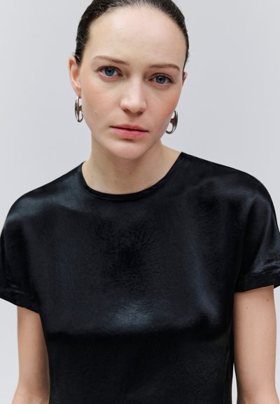 Junge Frau mit blauen Augen und zurückgebundenem Haar, trägt silberne Creolen und ein schwarzes Satin-Kurzarmoberteil, neutraler Gesichtsausdruck, hellgrauer Hintergrund.