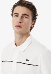 Valkoinen polo-paita, joka on valmistettu tekstuuri-kankaasta, jossa on vihreä brodeerattu krokotiililogo ja mustat vaakasuorat raidat, joissa on "LACOSTE"-teksti.