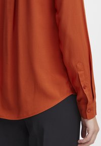 Chemise à manches longues en tissu orange rouille à la texture lisse, avec des poignets boutonnés et un ourlet arrondi, associée à un pantalon foncé.