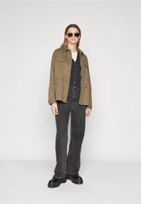 Khaki utility jacket with button details, layered over a dark vest and white shirt. Paired with black flared jeans and chunky black boots.