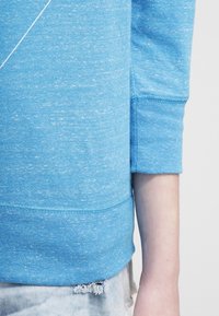 Ljusblå sweatshirt i strukturerad tyg med rund hals och ribbade ärmslut, med en horisontell söm vid fållen och lätt slitage.