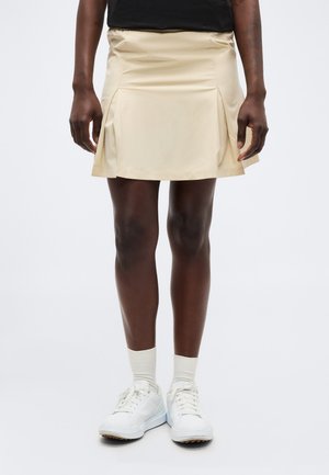 Sötét bőrű személy bézs pliszírozott szoknyát, fehér zoknit és fehér tornacipőt visel, egy egyszerű világos háttér előtt áll.