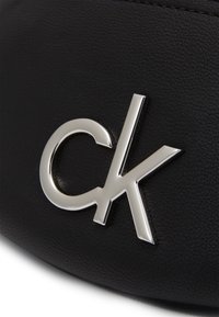 Černý materiál z umělé kůže s stříbrným logem "ck". Povrch má texturovaný vzhled a jemné detaily šití.