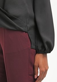 Blusa nera in raso con maniche a palloncino, abbinata a pantaloni aderenti bordeaux. Presenta uno spacco laterale sull'orlo per un dettaglio in più.