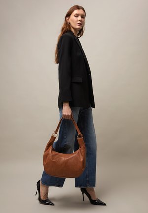 Vrouw in een zwart colbert en jeans, die een grote bruine leren handtas vasthoudt, staand in profiel tegen een effen achtergrond.