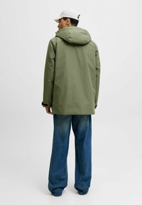 Grüne wasserdichte Jacke mit Kapuze, lockere Passform. Kombiniert mit blauen Jeans im weitem Schnitt. Die Jacke hat ein minimalistisches Design ohne sichtbare Hardware.
