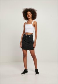 Witte cropped tanktop met vierkante halslijn gecombineerd met een zwarte denimrok. Model draagt zwarte high-top sneakers. Effen achtergrond.