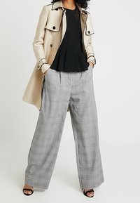 Trench-coat beige avec détails noirs, haut noir à basque et pantalon large à carreaux gris. Talons ouverts noirs complètent la tenue.