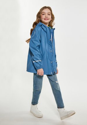 Giacca impermeabile blu con cappuccio, bottoni a pressione e dettagli in argento. Indossata sopra un paio di jeans denim e sneakers bianche. Tessuto liscio in tutto.