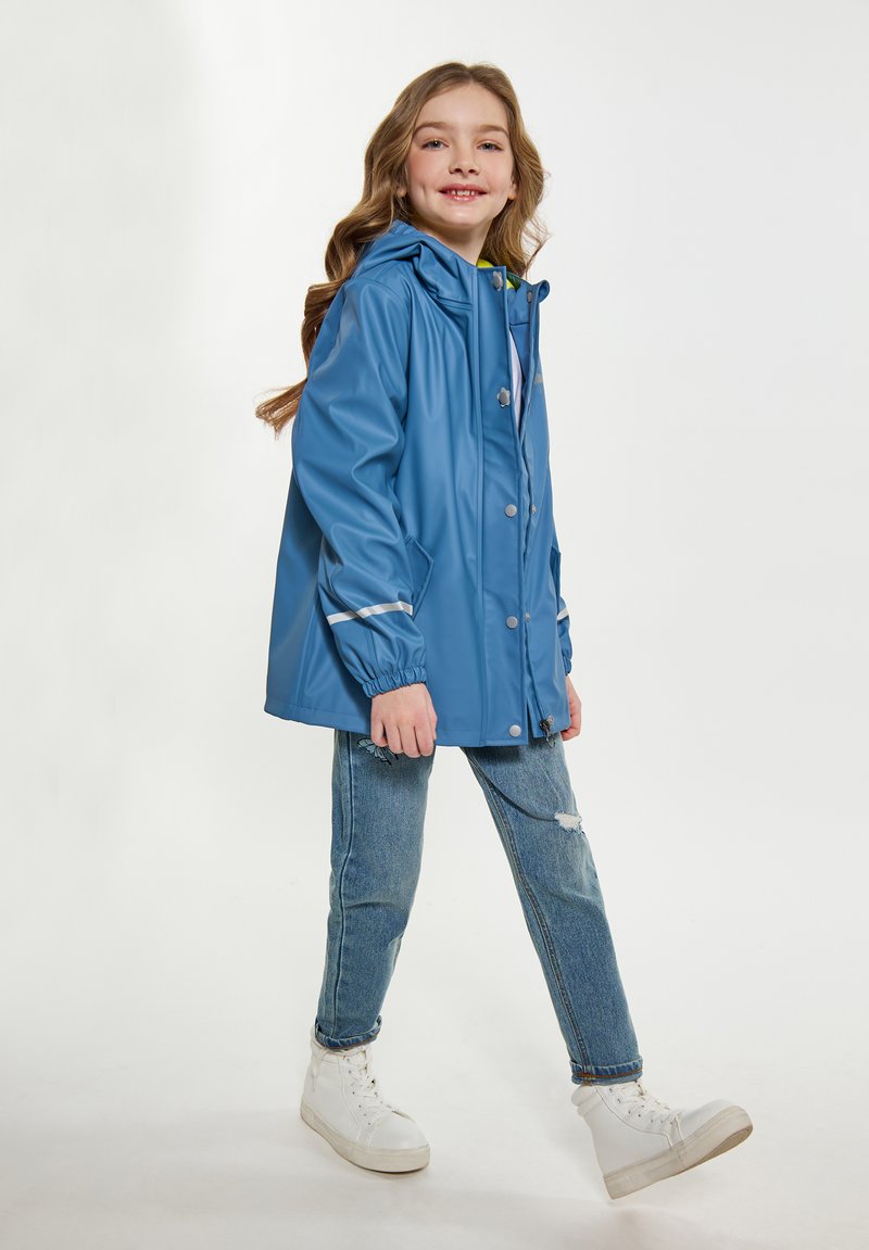 Blå vattentät jacka med huva, tryckknappar och silverdetaljer. Bärs över denimjeans och vita sneakers. Slät textur genom hela plagget.