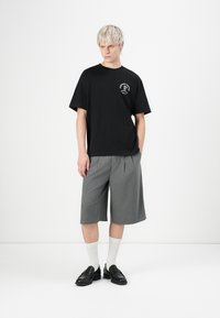Schwarzes kurzärmliges T-Shirt mit einem Logo, kombiniert mit grauen gefalteten Shorts. Getragen mit schwarzen Slippern und weißen Kniestrümpfen. Schlichter Hintergrund.