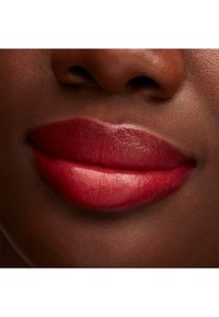Zbliżenie na pełne usta pokryte błyszczącą, ciemnoczerwoną szminką, podkreślające gładką teksturę i wyraźny kształt na tle ciemniejszego odcienia skóry.