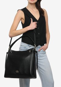Borsa a mano in pelle nera con forma strutturata, dotata di chiusura a cordoncino e dettagli metallici, portata a spalla con una lunga tracolla.