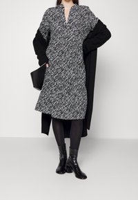 Schwarz-weiß gemustertes Kleid mit V-Ausschnitt, kurzen Ärmeln und ausgestelltem Saum, kombiniert mit einem langen schwarzen Cardigan und glänzenden Ankle Boots.