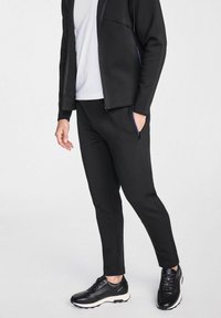 Czarne spodnie sportowe wykonane z gładkiego, elastycznego materiału z zwężanymi nogawkami i bocznymi kieszeniami na zamek. Połączone z czarnymi sneakersami i białą koszulą.