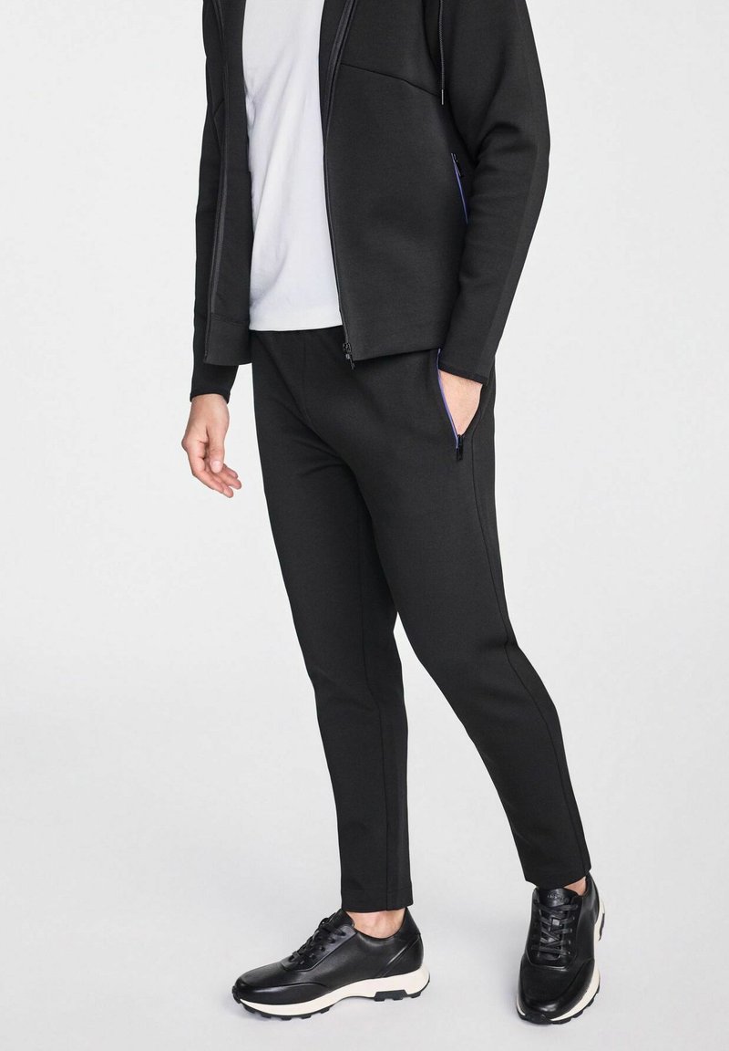 Czarne spodnie sportowe wykonane z gładkiego, elastycznego materiału z zwężanymi nogawkami i bocznymi kieszeniami na zamek. Połączone z czarnymi sneakersami i białą koszulą.
