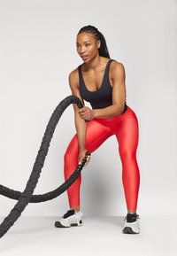 Fekete ujjatlan felső, piros fényes leggings és fehér sportcipő. Az egyén egy vastag fekete harci kötelet tart, edzés közben.