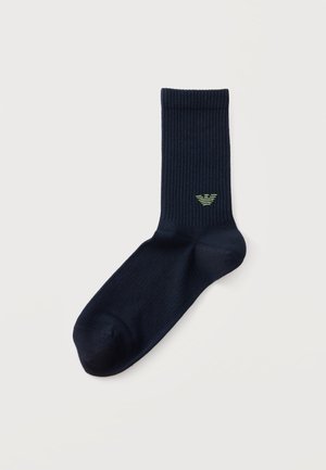 Une chaussette montante bleu marine foncé à côtes, ornée d’un petit logo vert clair, sur un fond blanc uni.