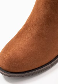 Stivale alla caviglia in suede marrone con punta arrotondata e superficie liscia. Presenta una suola scura a contrasto. La vista ravvicinata mette in risalto i dettagli del materiale.