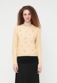 Femme aux cheveux roux bouclés portant un pull jaune pâle avec de petites fleurs brodées et une jupe noire, debout devant un fond blanc.