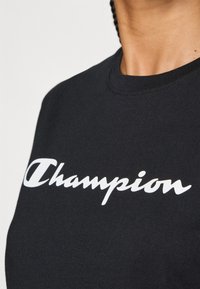 Crna pamučna majica sa dugim rukavima, koja nosi bijeli "Champion" logo u kurzivnom fontu, s klasičnim okruglim izrezom i mekanom teksturom.