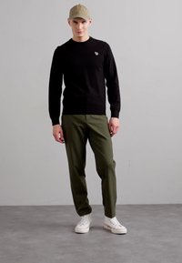 Μαύρο μακρυμάνικο μπλουζάκι με μικρό λογότυπο, πράσινο tapered παντελόνι και λευκά αθλητικά; το μοντέλο φοράει ένα ανοιχτό χακί καπέλο και στέκεται μπροστά σε γκρίζο φόντο.