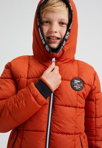 Oranssi puffatakki, jossa on tikattu muotoilu, musta vetoketju ja teksturoidut hihansuut. Siinä on huppu ja pyöreä logopatchi rinnassa.