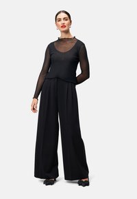 Haut noir à manches longues avec des panneaux transparents, associé à un pantalon noir à jambes larges. Le haut présente un décolleté arrondi et de subtils accents de volants.
