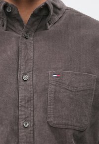 Γκρι πουκάμισο από κορντόρι με γιακά που κουμπώνει, τσέπη στο μπροστινό μέρος και διακριτικές κάθετες ρίγες. Διαθέτει μαύρα κουμπιά και μικρό λογότυπο.