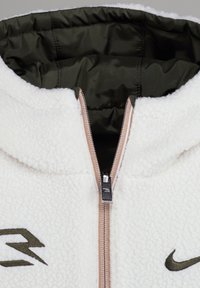 Veste à capuche en polaire blanche avec fermeture éclair, doublure intérieure vert olive et détails de logo brodés sur le devant.