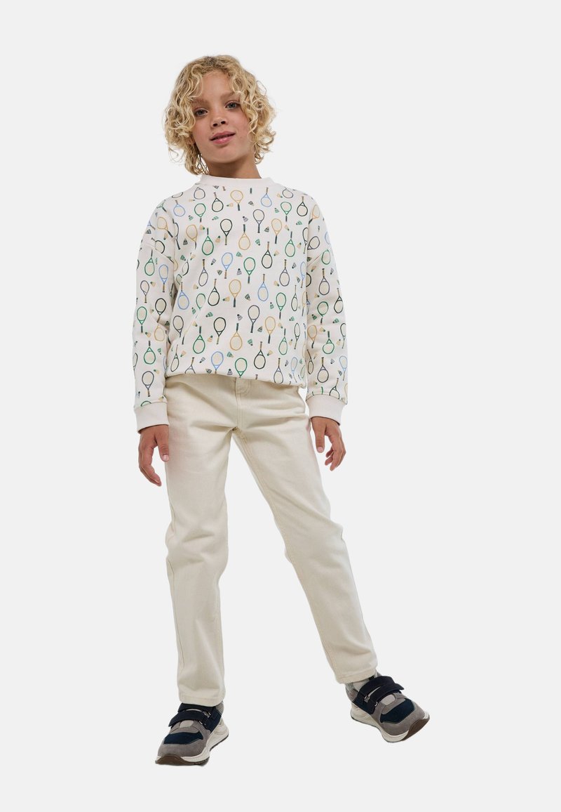 Sweatshirt blanc avec des motifs de raquettes de tennis multicolores, associé à un pantalon de couleur crème. Enfant portant des baskets noires et grises.