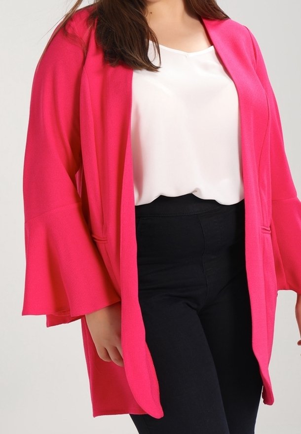 Mujer con chaqueta abierta de color rosa brillante con mangas anchas, blusa blanca y pantalones negros ajustados sobre un fondo liso.