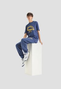 Tuta sportiva blu con testo giallo, realizzata in tessuto morbido, caratterizzata da una vestibilità rilassata. Sneakers bianche con dettagli blu. Modello seduto su un piedistallo.