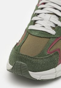 Baskets vertes et marron avec textures en maille et en daim, lacets blancs et accents roses subtils ; disposent d'une semelle texturée et d'un col rembourré.