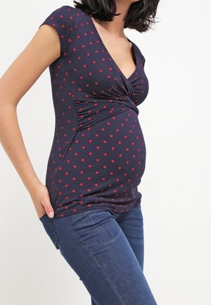 Zwangere vrouw draagt een marineblauw, kort mouwtje, rood met witte stippen top en blauwe spijkerbroek, met één hand in haar zak.