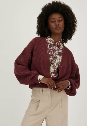 Bordeaux gebreide cardigan met een diepe V-hals, oversized mouwen en een enkele knopen-sluiting, gelaagd over een gebloemde groene en bordeaux blouse.