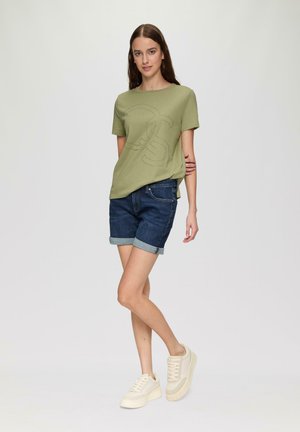 T-shirt à manches courtes vert olive avec un logo en relief discret, associé à un short en jean roulé et des baskets de couleur claire.