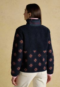 Marineblauwe fleece trui met een hoge kraag, voorzien van een bloemenpatroon in bordeauxrood en roze, en een logo op de kraag. Zachte, gestructureerde stof.