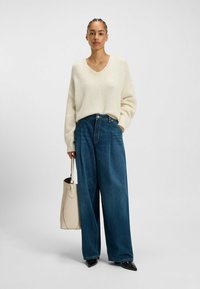 Sweterek w kolorze kremowym, luźny, z głębokim dekoltem w serek, połączony z niebieskimi dżinsami o wysokim talii i szerokich nogawkach oraz jasnobeżową torbą tote.