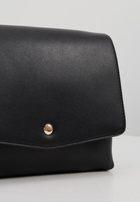 Černá kožená kabelka s uzávěrem na zlaté tlačítko na bílé ploše a světle šedém pozadí.