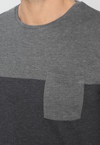 Camiseta gris con un escote redondeado, con un diseño bicolor: gris claro en la parte superior y gris oscuro en la parte inferior, y un bolsillo en el pecho.
